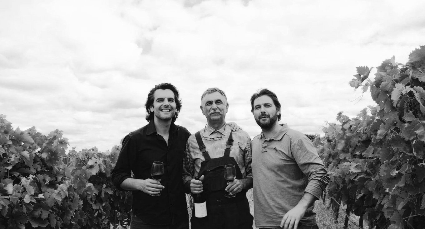 Três homens em um vinhedo, segurando taças e uma garrafa de vinho.